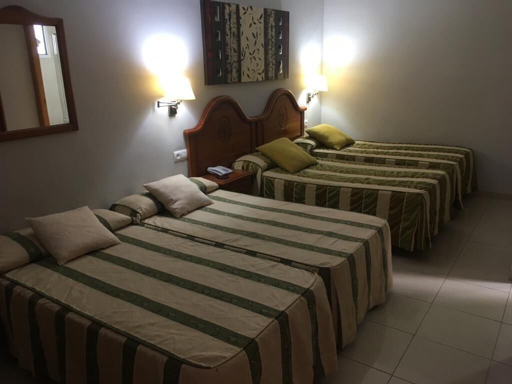 A hotel room with four beds arranged in a row, each with a headboard and pillows. There are wall-mounted lamps on either side of the beds.
