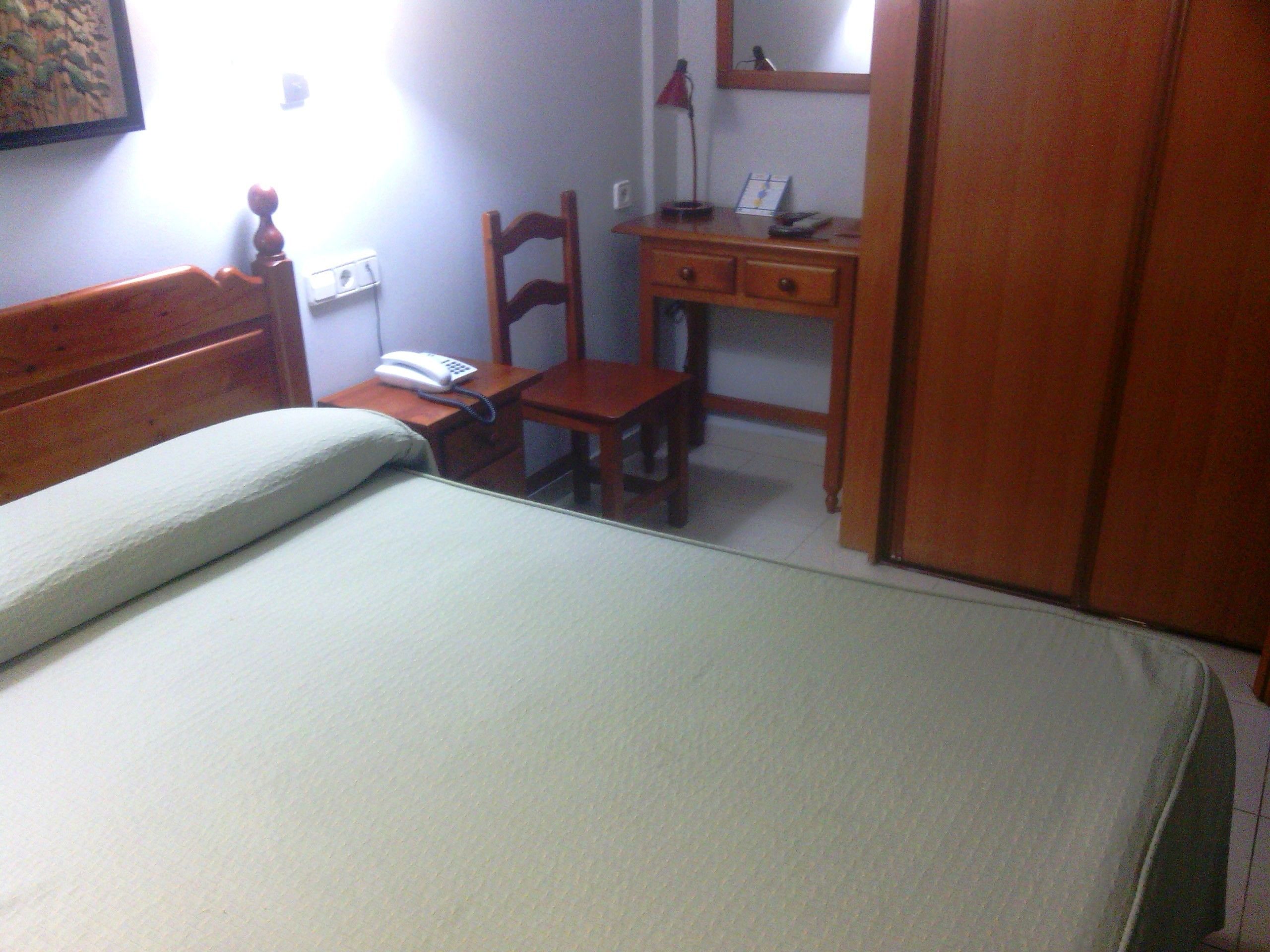 Un dormitorio con una cama, una mesita de noche de madera con un teléfono, un escritorio con una lámpara y una silla.
