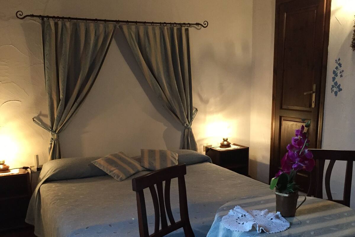 A bedroom with a bed, a chair, a nightstand, a lamp, and a potted plant.