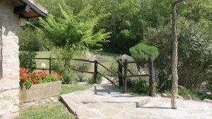 Un sendero de piedra que conduce a una estructura de madera rústica con una puerta de metal, rodeada de abundante vegetación y un muro de piedra con flores rojas.