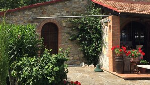 Un edificio de piedra con techo de tejas rojas, una chimenea y un ventanal pequeño. Hay macetas con flores rojas y un camino de piedra que lleva a la entrada.