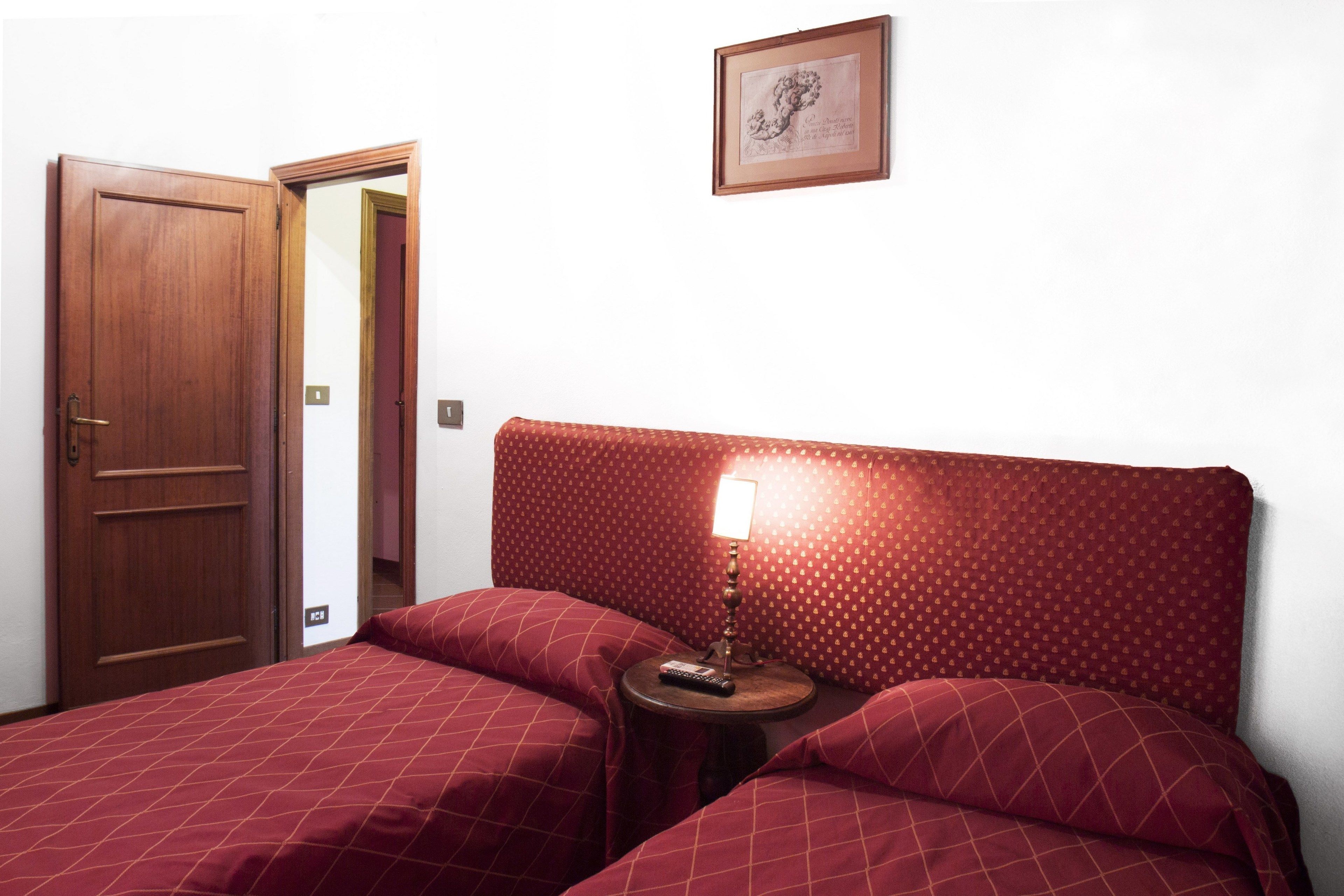 Una habitación con puerta de madera, cama con colcha estampada roja, mesita de noche con lámpara y un cuadro en la pared.