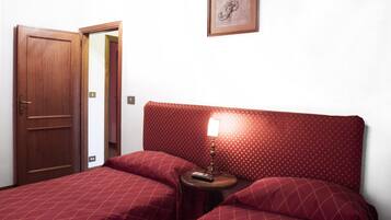 Una habitación con puerta de madera, cama con colcha estampada roja, mesita de noche con lámpara y un cuadro en la pared.