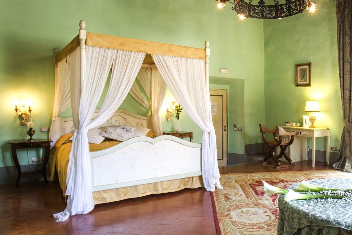 Un lit à baldaquin avec des rideaux blancs, une tête de lit en bois et une couvre-lit à motifs floraux. Un petit guéridon avec une lampe et une chaise se trouvent dans un coin. Les murs de la pièce sont peints en vert et un tapis à motifs recouvre le sol.