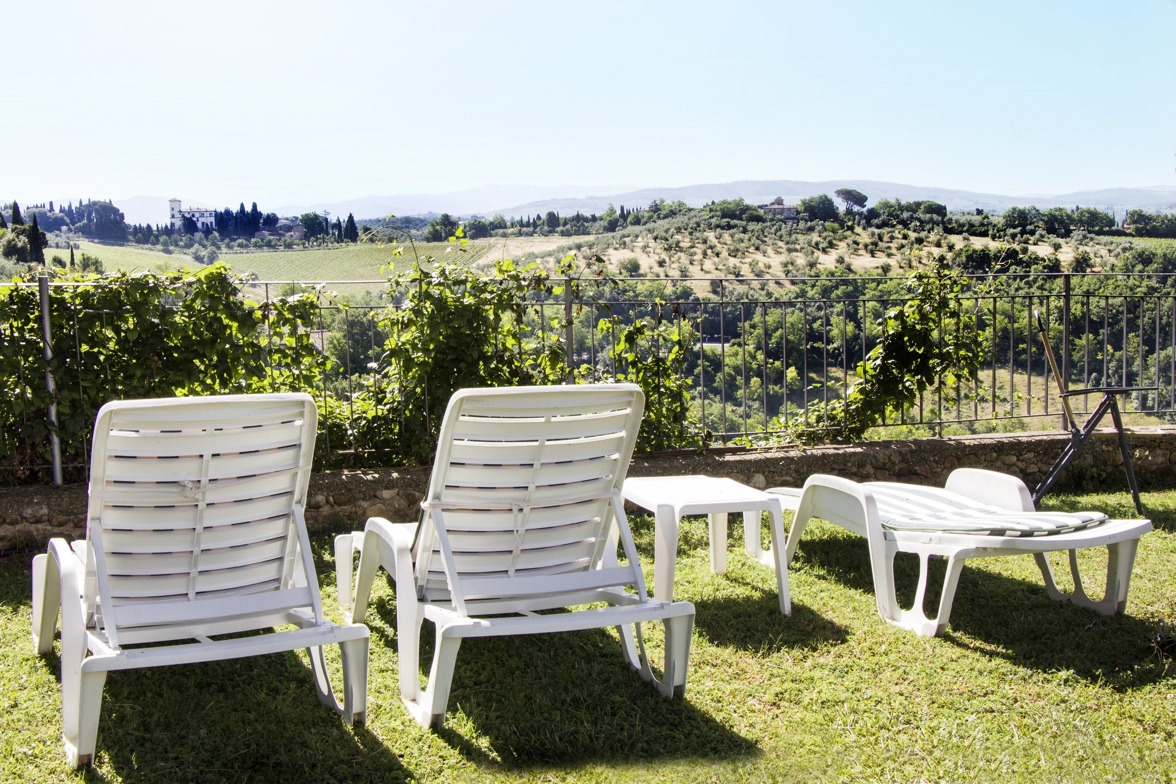 Three white plastic lounge chairs and a small table on a grassy lawn with a vineyard and distant hills in the background.