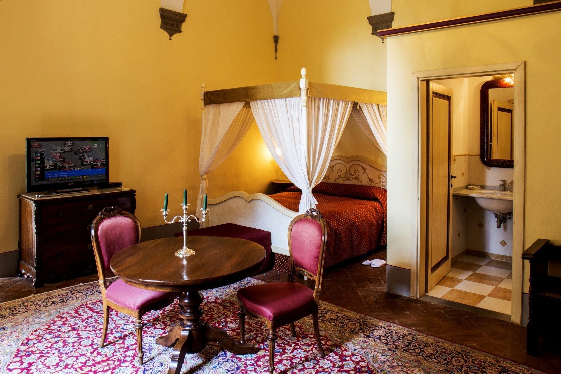 A room with a four-poster bed, a bedside table with a candelabra, a television on a wooden cabinet, and a checkered floor.
