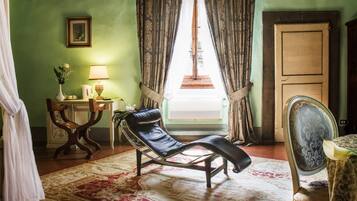 A room with a chaise lounge, a wooden chair, a table with a lamp, a picture on the wall, and a window with curtains.