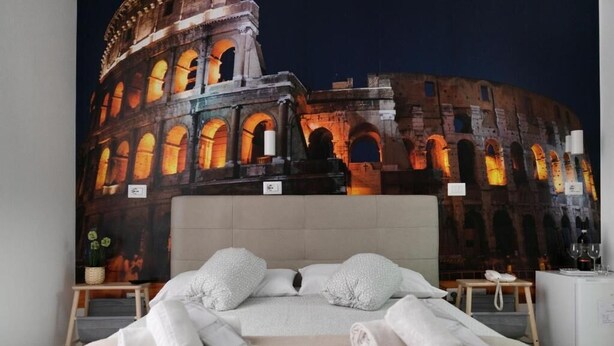 A hotel room with a large bed, a nightstand, a small table with a lamp, and a wall-mounted TV displaying a nighttime view of the Colosseum. - Merulana Star (Rome)