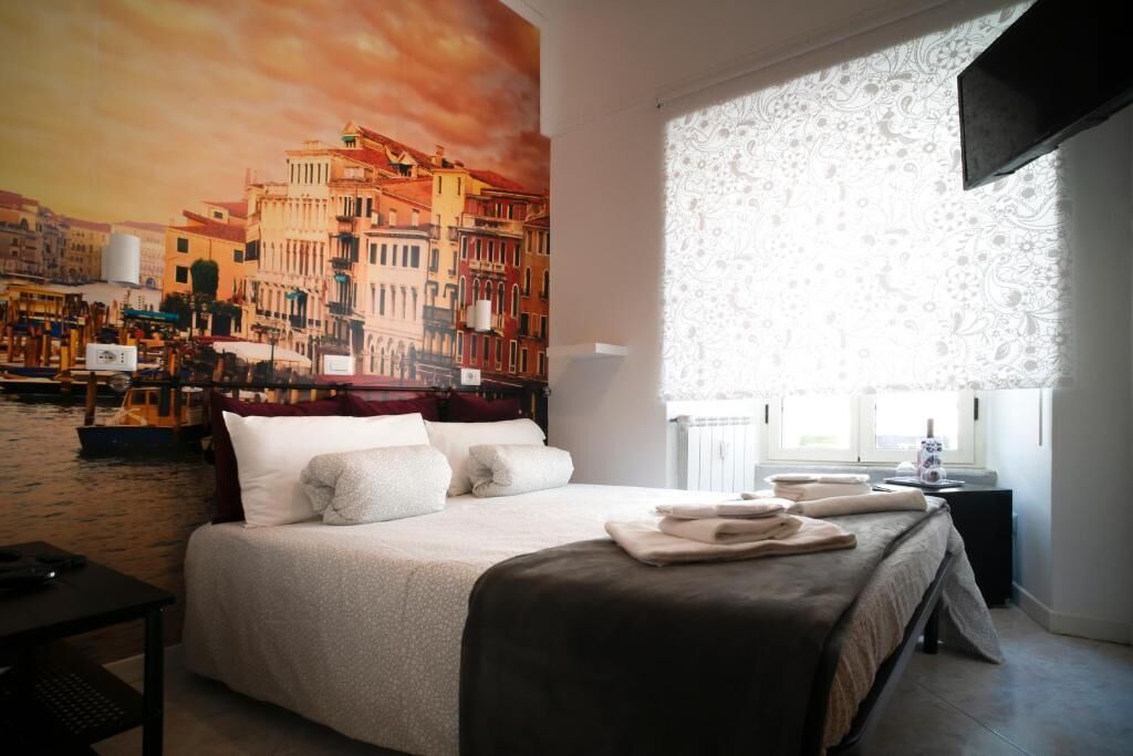 Ein Hotelzimmer mit einem großen Bett, einem Stadtbild als Gemälde, einem Fenster mit gemusterten Vorhängen und einem Nachttisch mit Lampe.