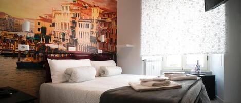 Ein Hotelzimmer mit einem großen Bett, einem Stadtbild als Gemälde, einem Fenster mit gemusterten Vorhängen und einem Nachttisch mit Lampe.