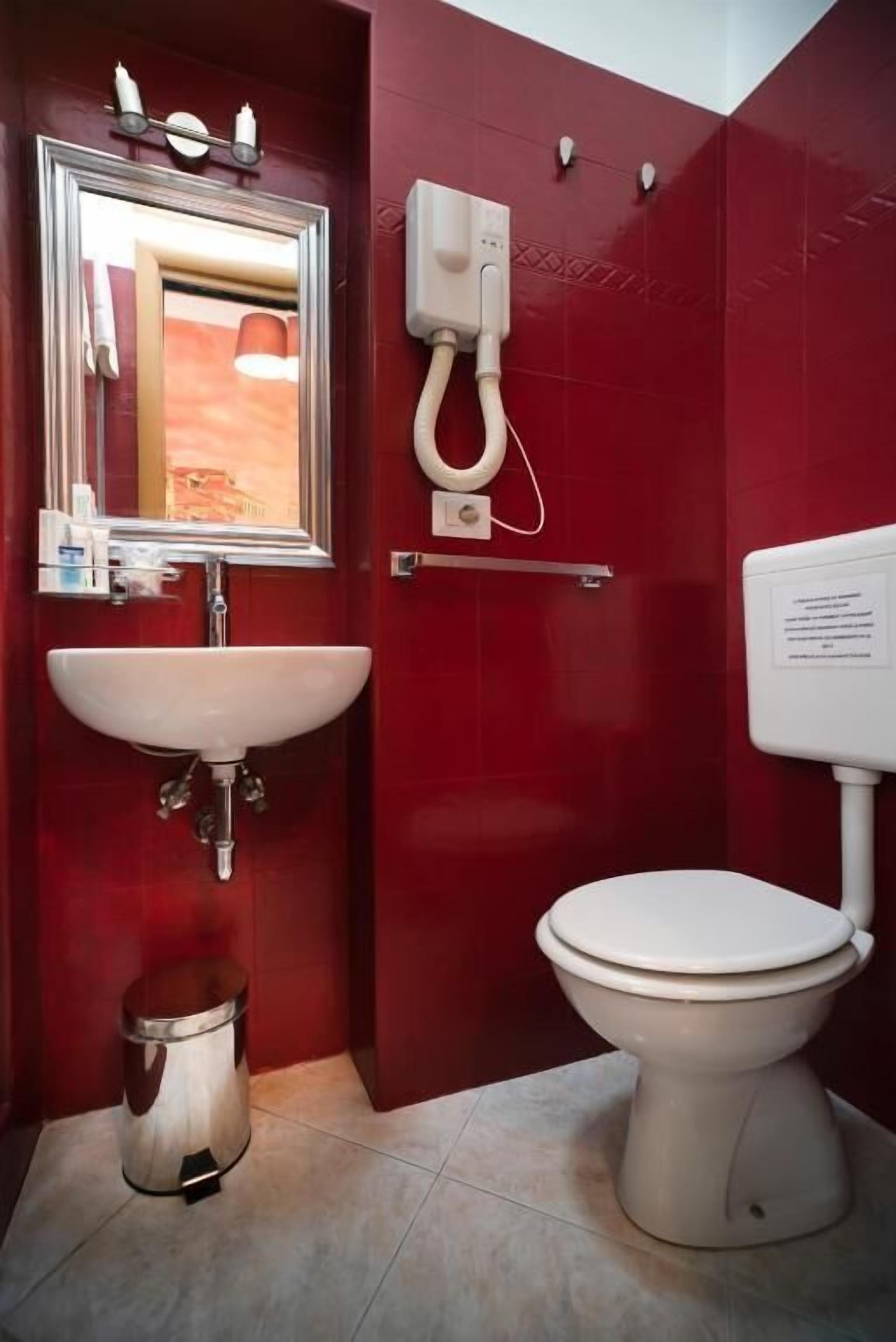 A bathroom with a red tiled wall, a white sink, a mirror, a toilet, and a trash can.