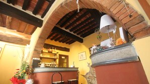 Um espaço de bar acolhedor com teto de tijolos rústicos, uma coluna de pedra e uma luminária pendente. Há dois bancos e uma mesa pequena com um vaso vermelho e um castiçal branco.