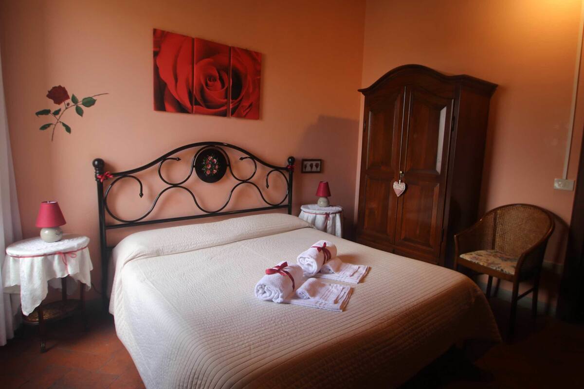 A neatly made bed with rolled towels, a wooden wardrobe, a wicker chair, and a small table with a lamp.