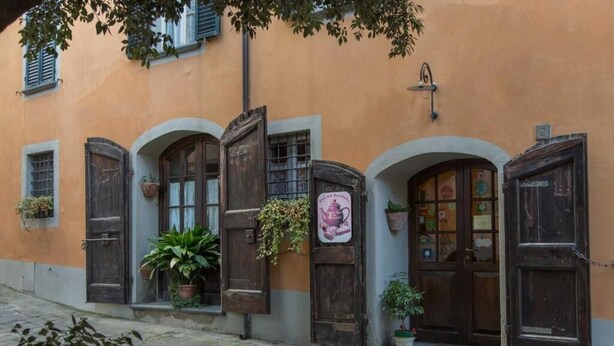 Um edifício com portas e janelas em arco, algumas com persianas. Há plantas em vasos e uma placa na parede.