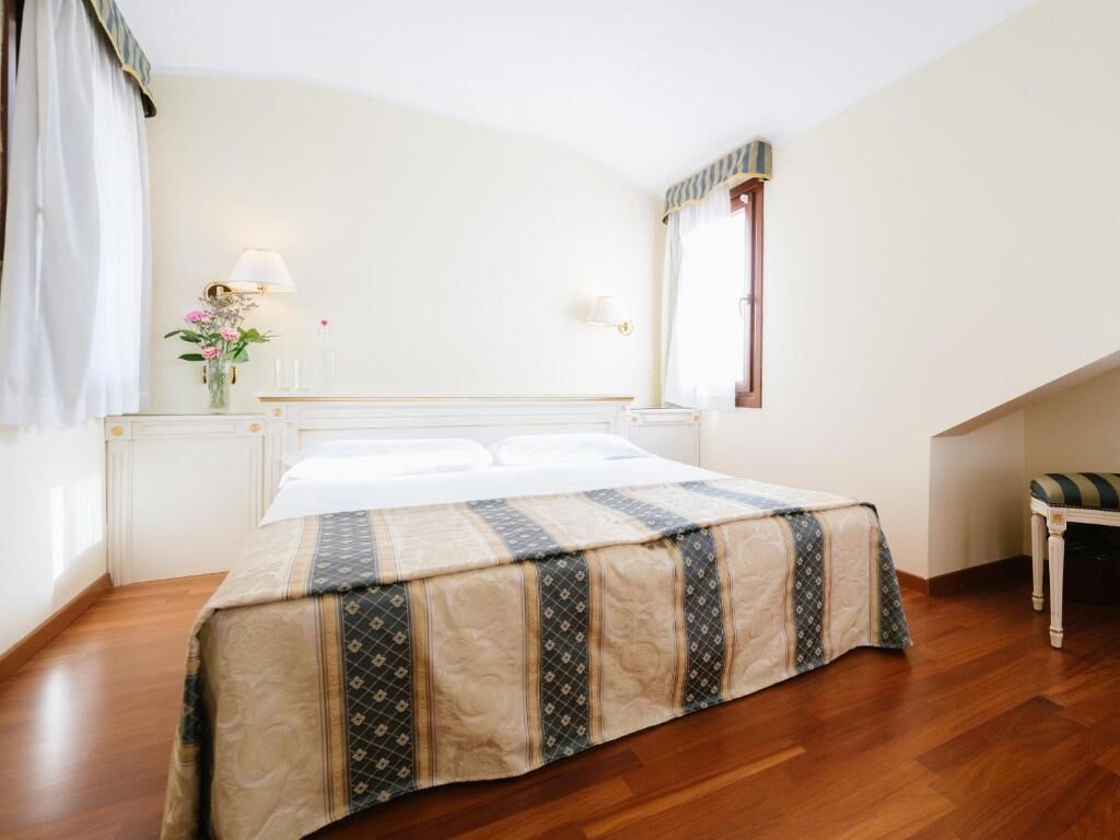 A neatly made bed with a patterned bedspread, a bedside table with a lamp, and a window with curtains.