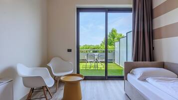 Ein modernes Hotelzimmer mit einem Bett, zwei weißen Stühlen, einem kleinen Tisch und einer Schiebetür zu einem Garten.