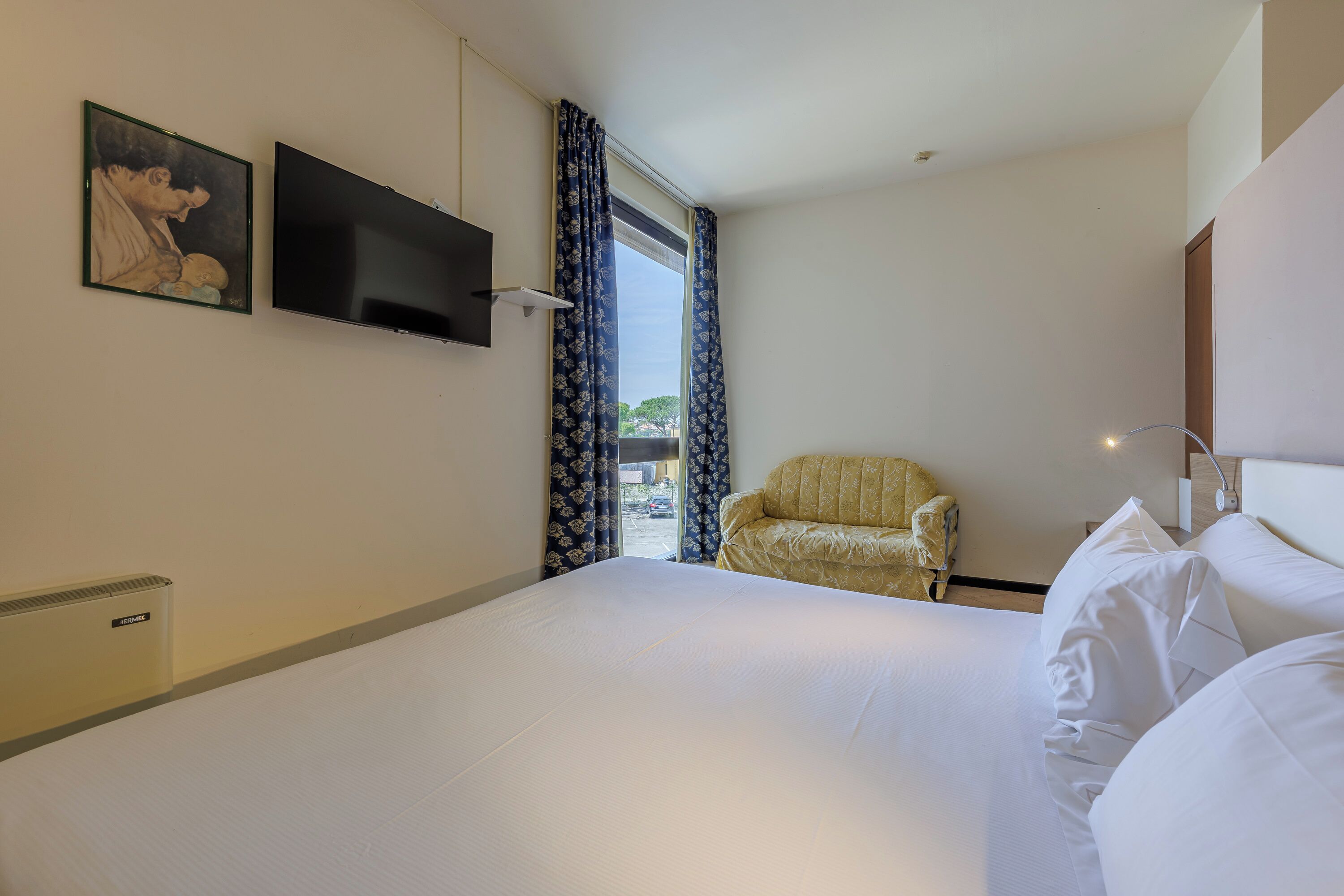 Quarto de hotel com uma cama, uma cadeira, uma televisão e uma janela com cortinas.