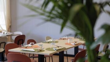 A dining area with tables set for four, chairs, and a large plant in the foreground.