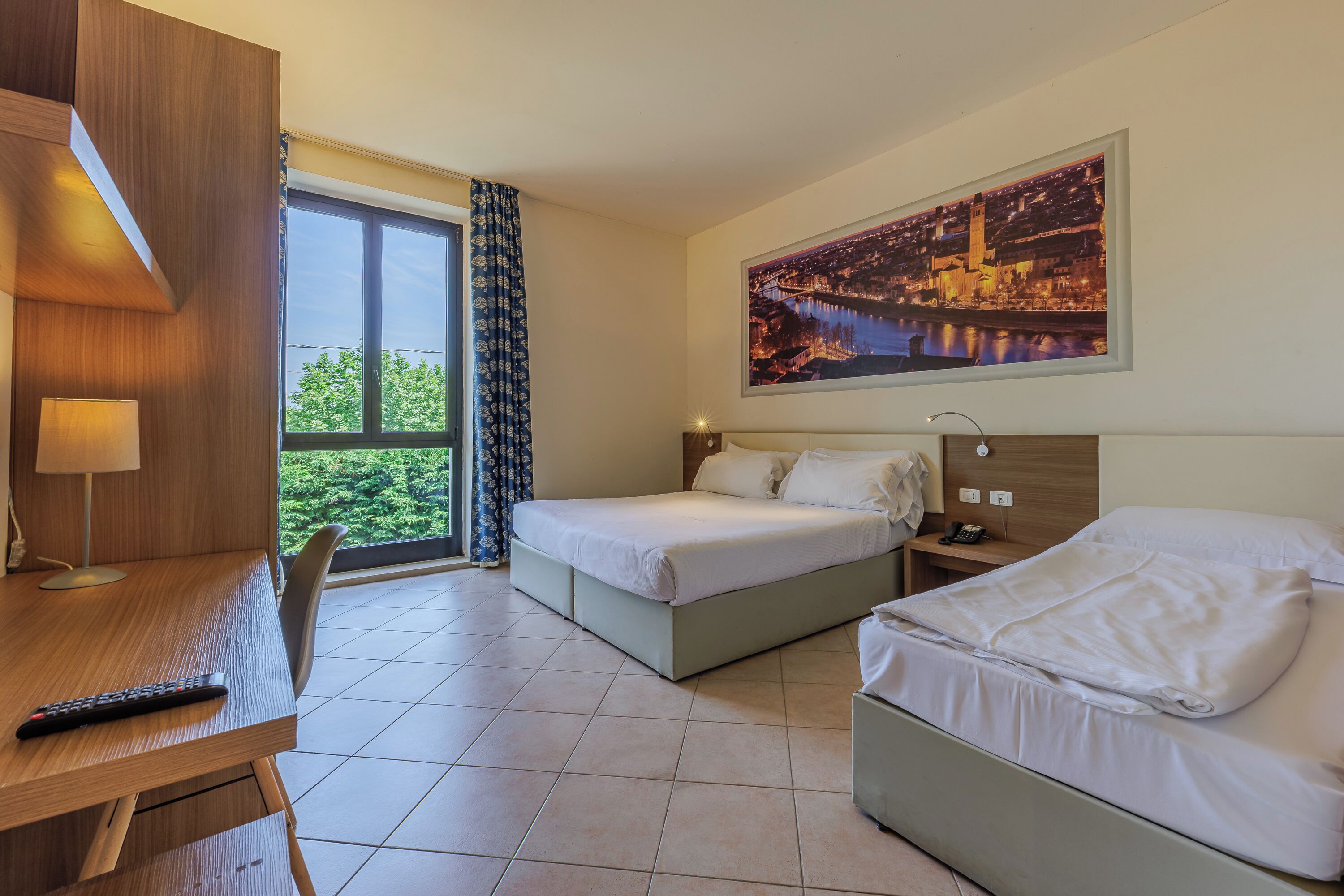 A hotel room with two beds, a desk, a chair, a window with curtains, a painting, and a lamp.
