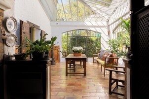 Un invernadero con una mesa y sillas de madera, rodeado de plantas en macetas y vistas a zonas verdes.
