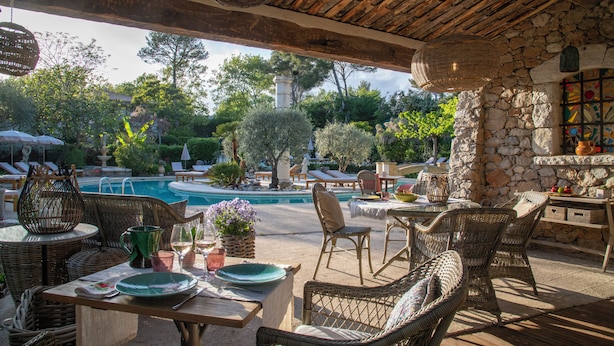 An outdoor patio with wicker furniture, a stone wall, and a view of a swimming pool and garden. - Hotel Les Messugues (Saint-Paul-de-Vence)