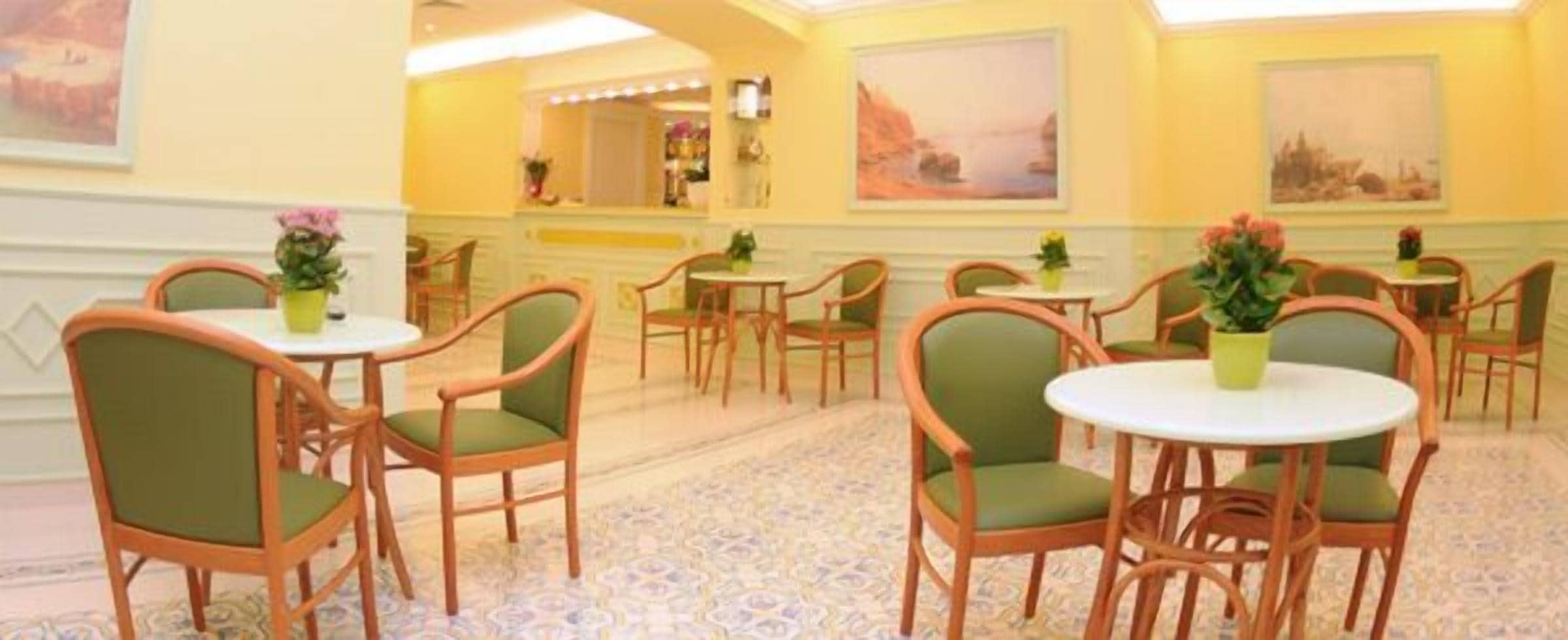 A hotel lobby with multiple round tables and green upholstered chairs, a patterned carpet, and artwork on the walls.