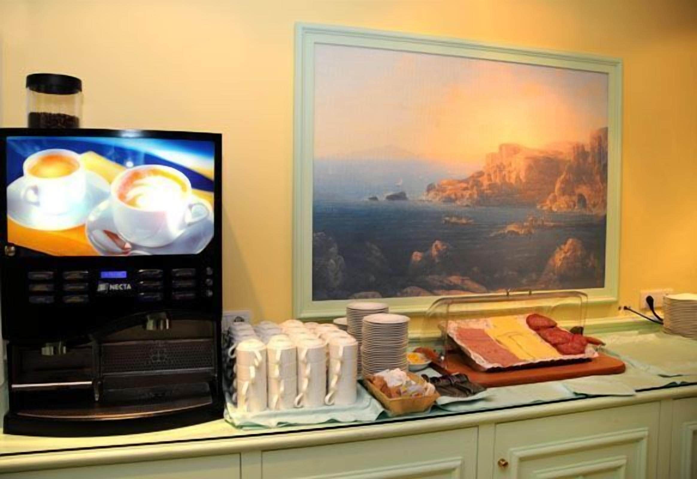 A coffee machine, plates, and food on a counter with a painting of a coastal scene in the background.