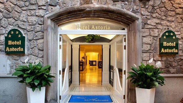 La entrada del Hotel Astoria, con fachada de piedra, un felpudo azul en la entrada y plantas en macetas a cada lado.