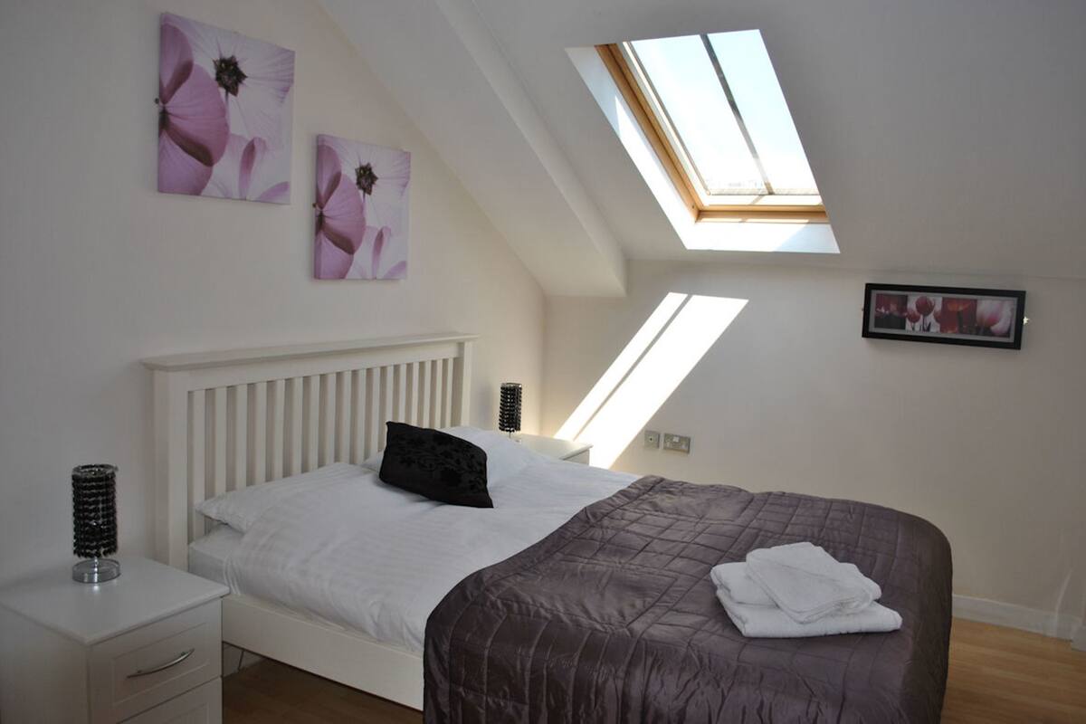 A bedroom with a sloped ceiling, skylight, white bed, dark brown bedspread, white bedside tables, and wall art.