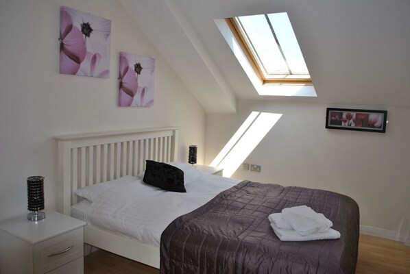 A bedroom with a sloped ceiling, skylight, white bed, dark brown bedspread, white bedside tables, and wall art.
