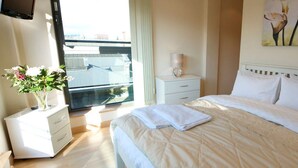 A hotel room with a bed, a bedside table, a nightstand, a window with a view, and a painting on the wall. - Dreamhouse Manchester Deansgate (Manchester)