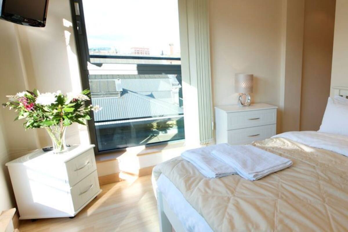 A hotel room with a bed, a bedside table, a nightstand, a window with a view, and a painting on the wall.