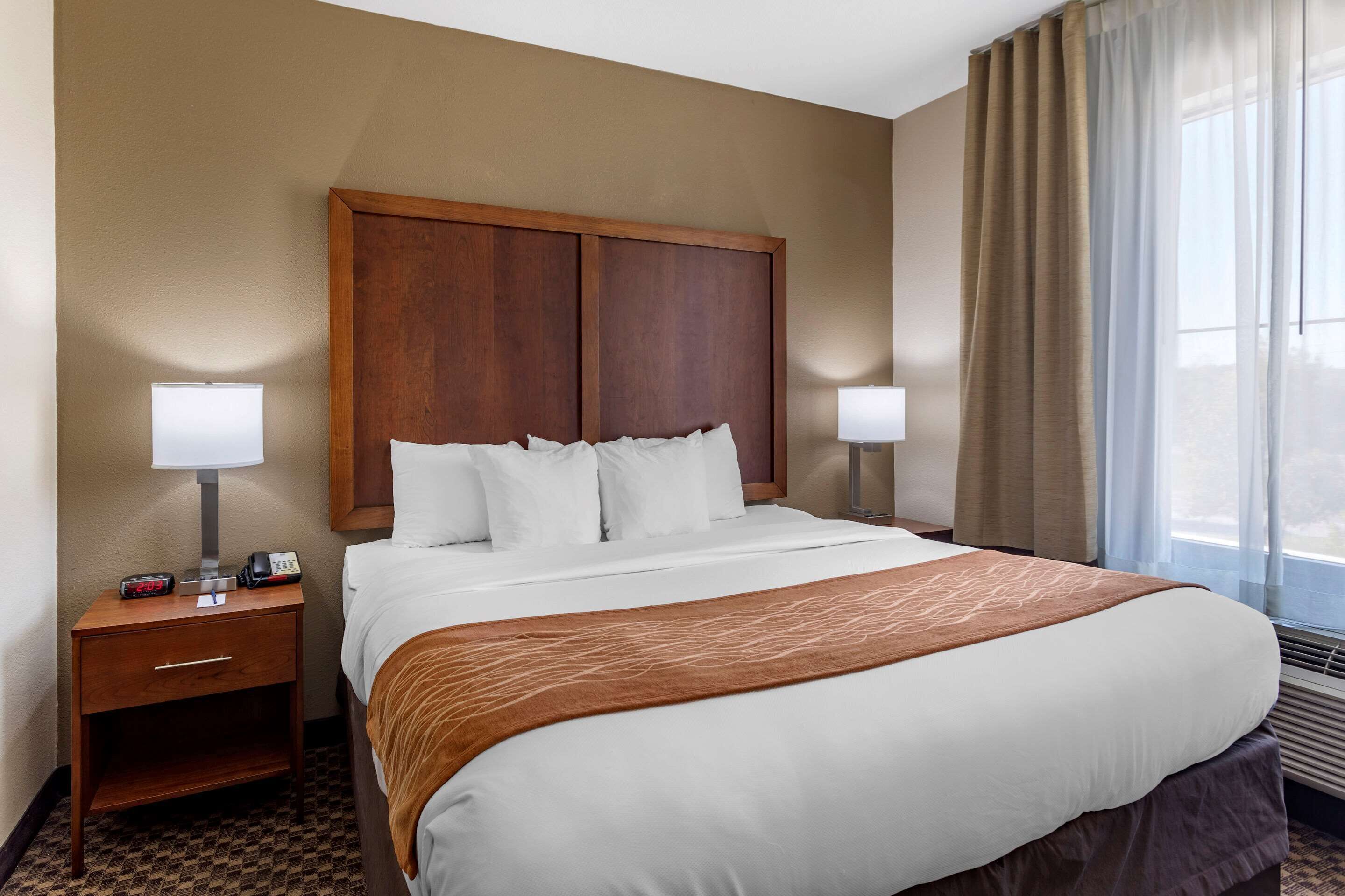 Une chambre d’hôtel avec un grand lit, deux tables de chevet avec des lampes, une tête de lit en bois et une fenêtre avec des rideaux.