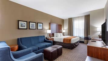A hotel room with a bed, a blue sofa, a wooden side table, a desk with a television, and framed pictures on the wall.