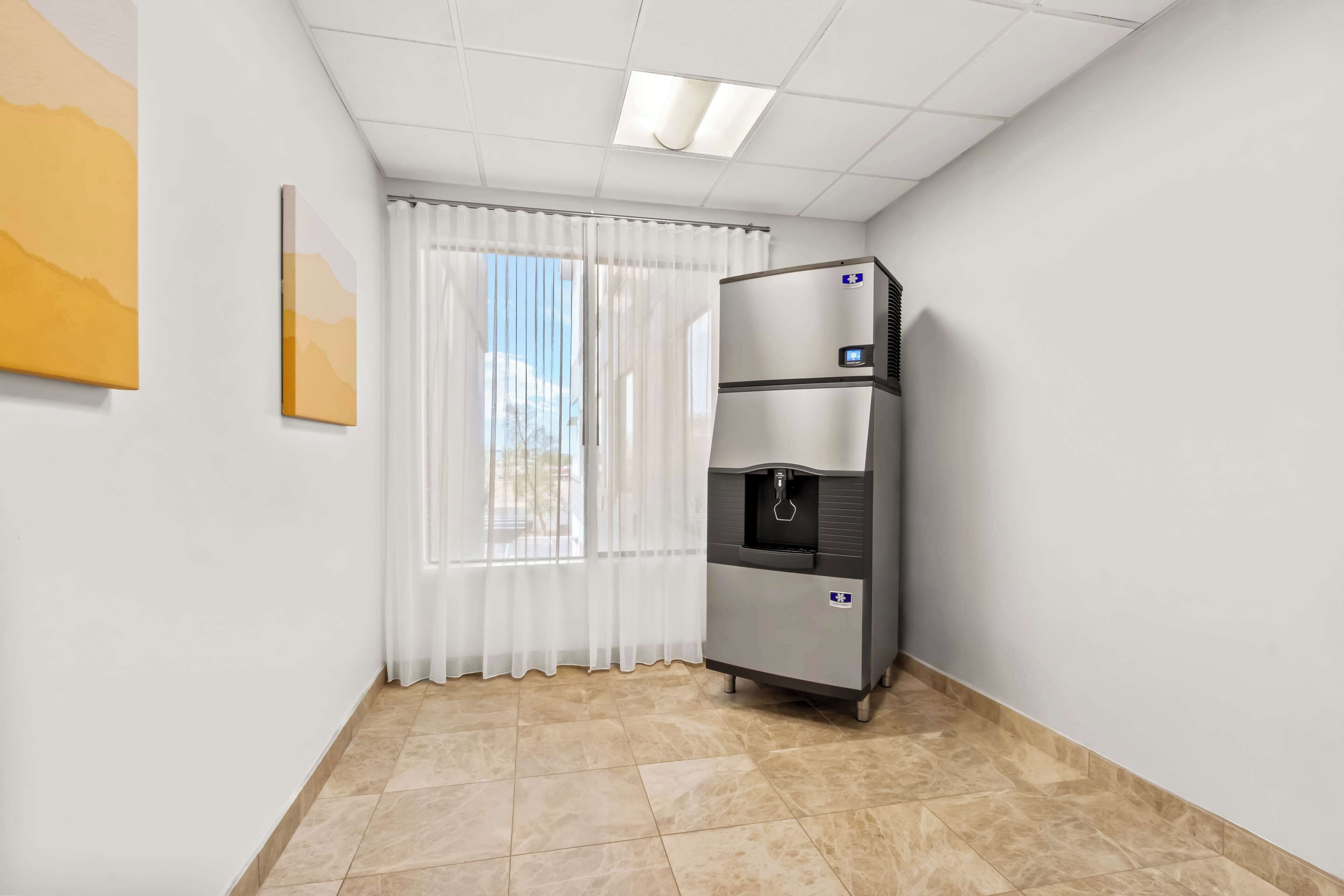 A hotel room with a water dispenser, a window with white curtains, and a painting on the wall.