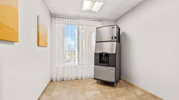 A hotel room with a water dispenser, a window with white curtains, and a painting on the wall.
