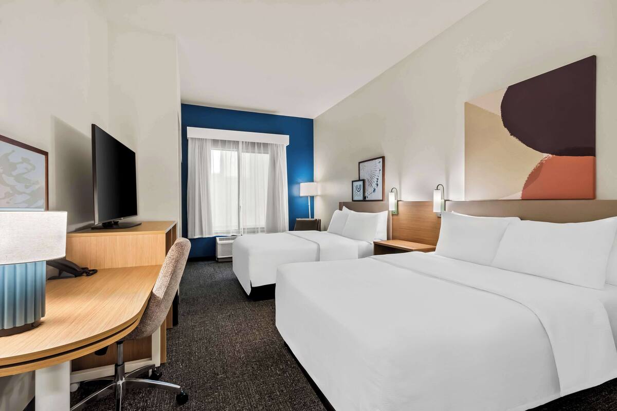 A hotel room with a desk, two beds, a television, a window with curtains, and a painting on the wall.