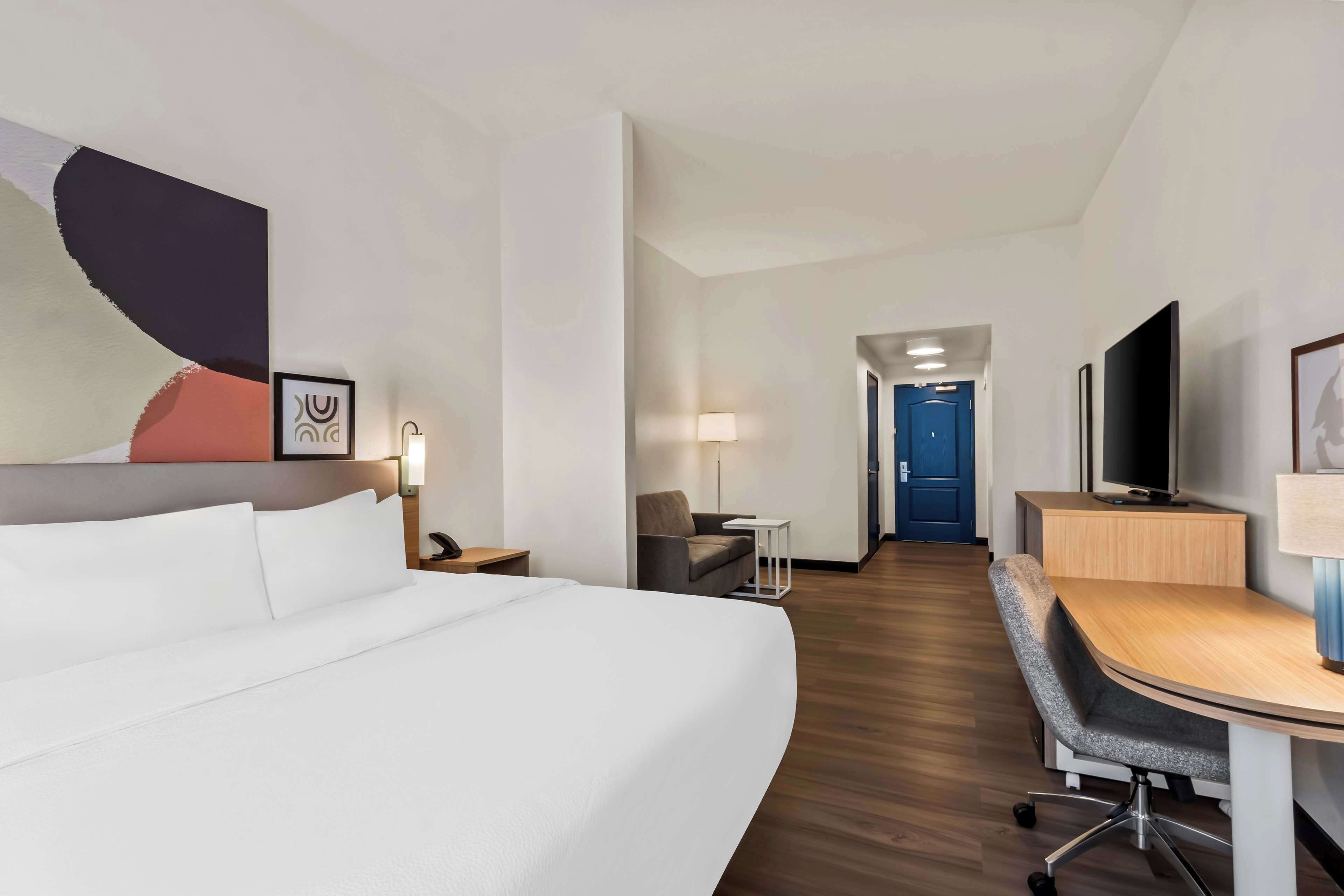 A hotel room with a large bed, a desk with a chair, a television, and a door leading to another room.
