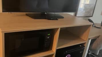 A compact kitchenette with a microwave, a small refrigerator, and a flat-screen TV on a wooden desk.