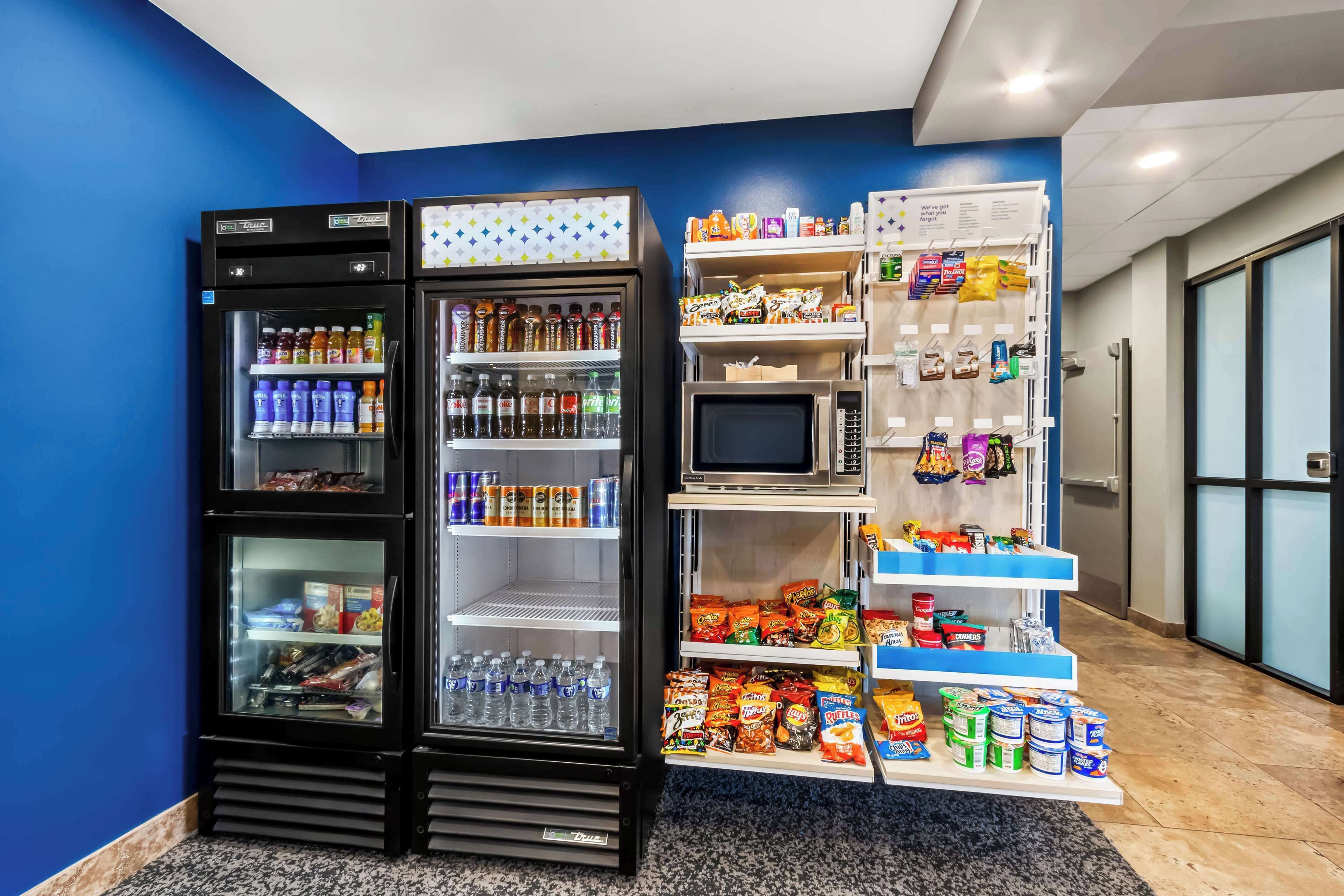 A vending machine with a variety of drinks and snacks, a microwave, and a shelf stocked with packaged food items.