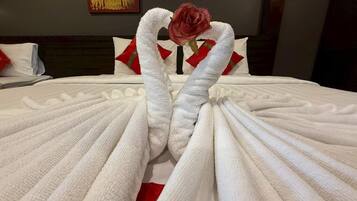 Uma cama bem arrumada com toalhas dispostas em formato de coração, uma rosa solitária e uma almofada decorativa com padrão vermelho e dourado.