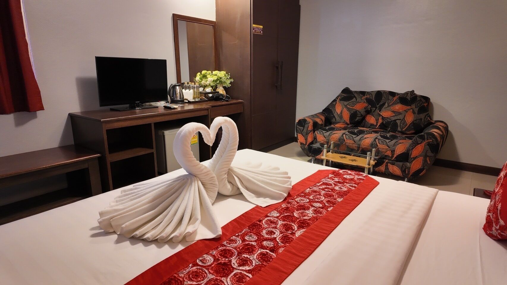 Habitación de hotel con una cama decorada con un cisne de toallas, un corredor floral rojo, un televisor sobre un escritorio y un sillón estampado.