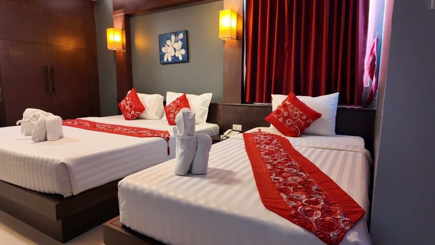 A hotel room with two beds, each with white linens and red decorative pillows. There are towels placed on the beds, and a red decorative runner is draped over the beds.