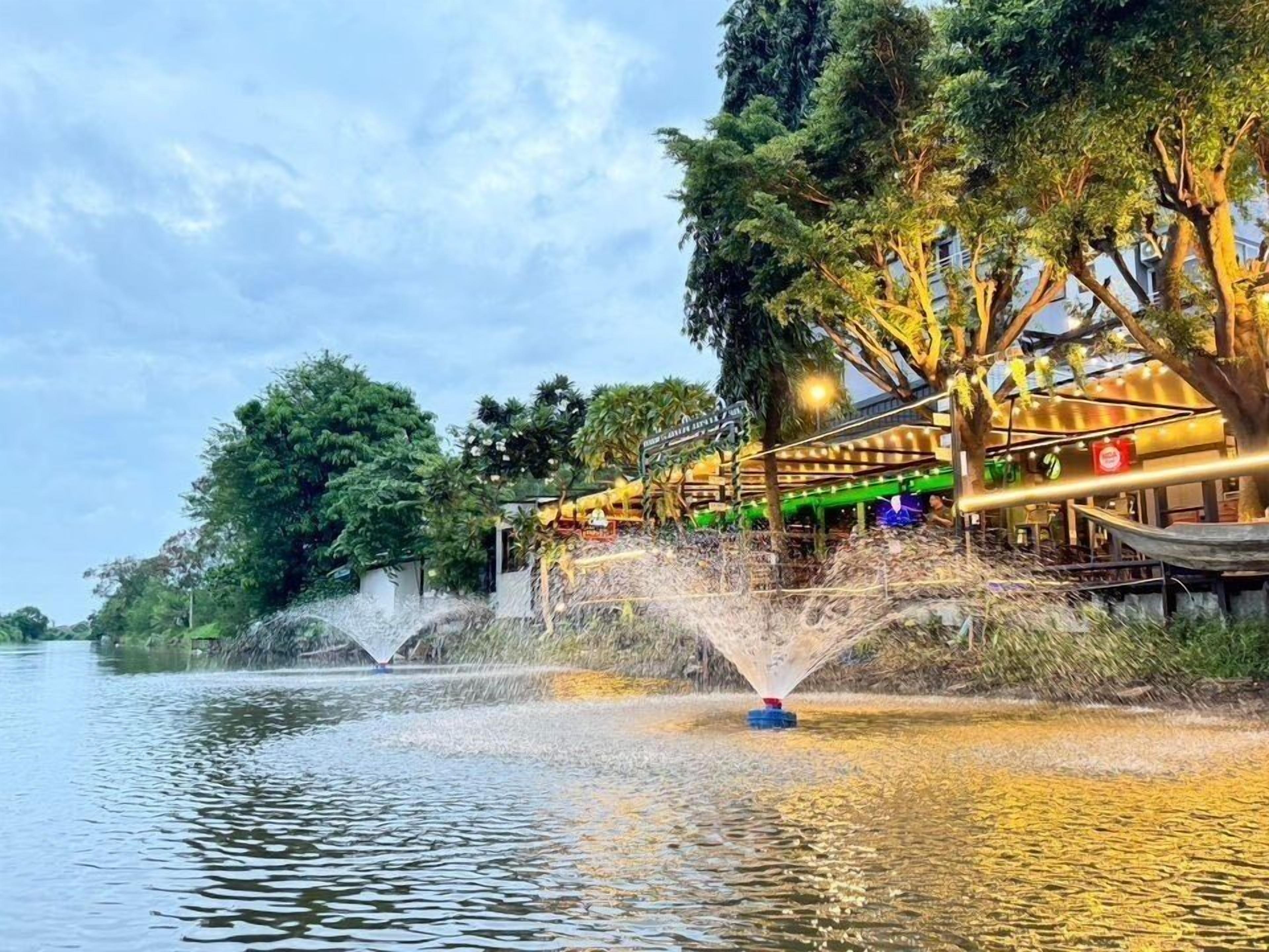 A riverside area with a water fountain, trees, and a building with outdoor seating.