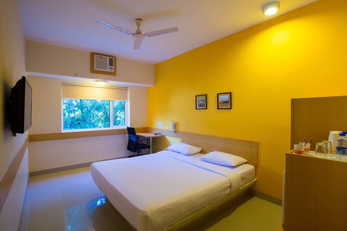 A hotel room with a bed, a desk, a chair, a ceiling fan, and a window with a view of trees.