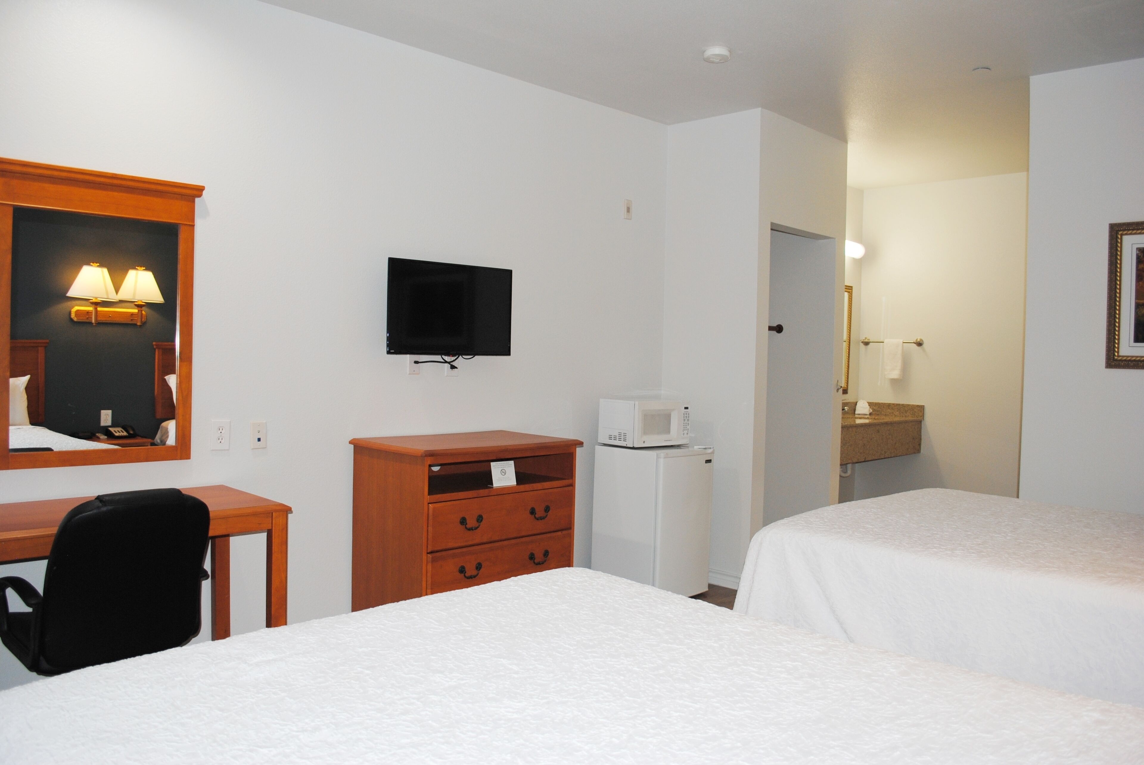 Habitación de hotel con dos camas, un escritorio, una silla, un televisor, un microondas y un baño pequeño con espejo y toallero.
