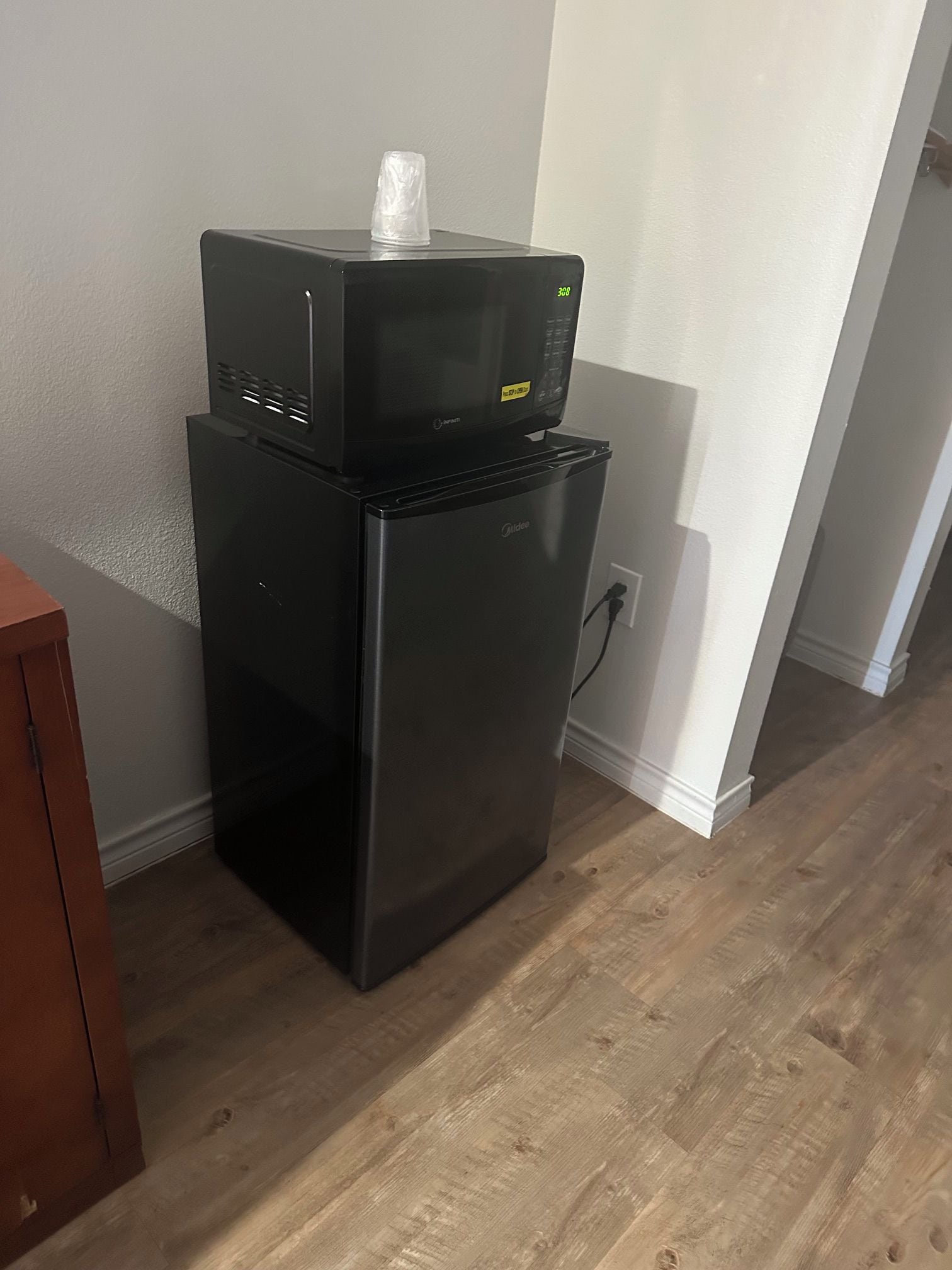 Un refrigerador negro con un microondas encima, un enchufe en la pared y piso de madera.