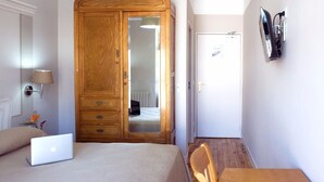 A bedroom with a bed, a laptop, a wooden wardrobe, a mirror, a wall-mounted TV, and a painting. - Majestic (Lourdes)