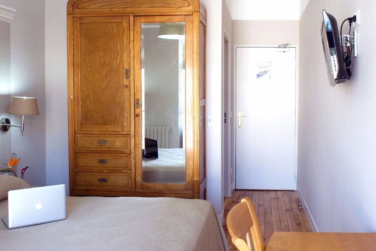 A bedroom with a bed, a laptop, a wooden wardrobe, a mirror, a wall-mounted TV, and a painting.