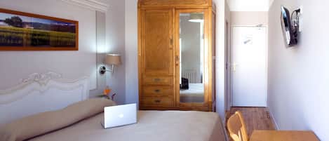 Un dormitorio con cama, laptop, armario de madera, espejo, televisor montado en la pared y un cuadro.